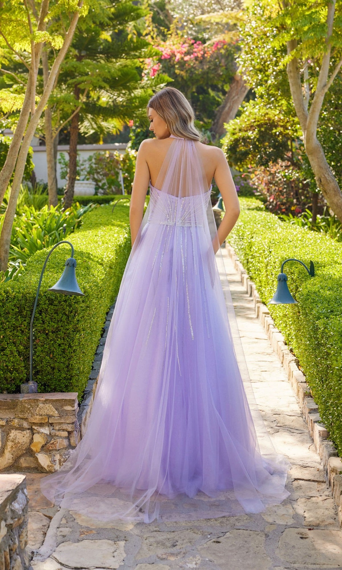 Wearing the Long Prom Dress Juliet JT2620U with a rhinestone bodice and sheer cape, a woman stands on a sunlit garden path, surrounded by vibrant greenery and flowers.